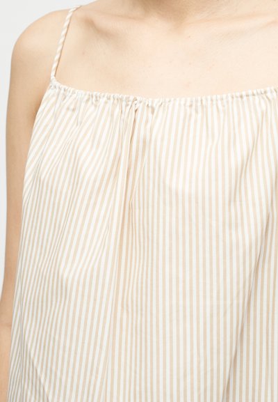 Κοντινό πλάνο ενός ατόμου που φοράει αμάνικο τοπ με κάθετες ρίγες σε μπεζ και άσπρο, με λεπτές τιράντες σαν σπάγγο.