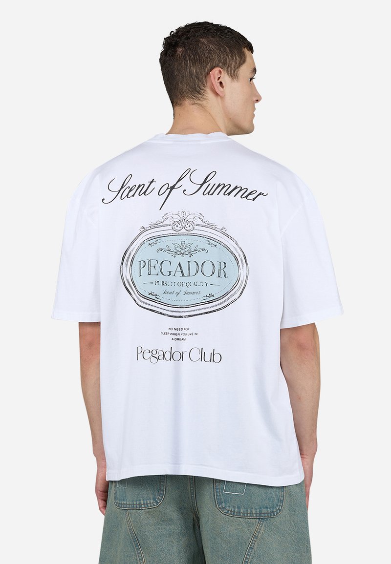 Fehér pamut póló, kék ovális grafikával, amelyen a "PEGADOR" és a "Nyár Illata" felirat szerepel, rövid ujjakkal, laza szabással és kerek nyakkivágással.