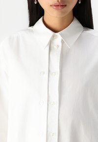 White button-up shirt made of a smooth fabric, featuring a classic collar, and translucent buttons, with a relaxed fit and no visible patterns.