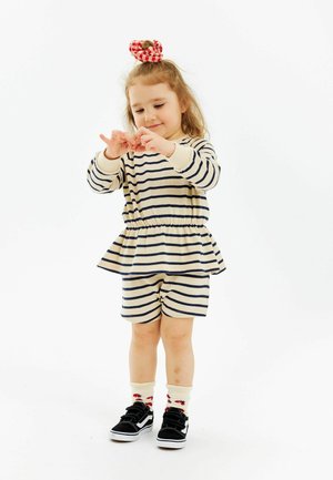 Randig beige och marinblå outfit med en peplumtopp och shorts. Barnet har svarta sneakers och krämfärgade strumpor med blomdekorationer, håret uppsatt med en röd scrunchie.