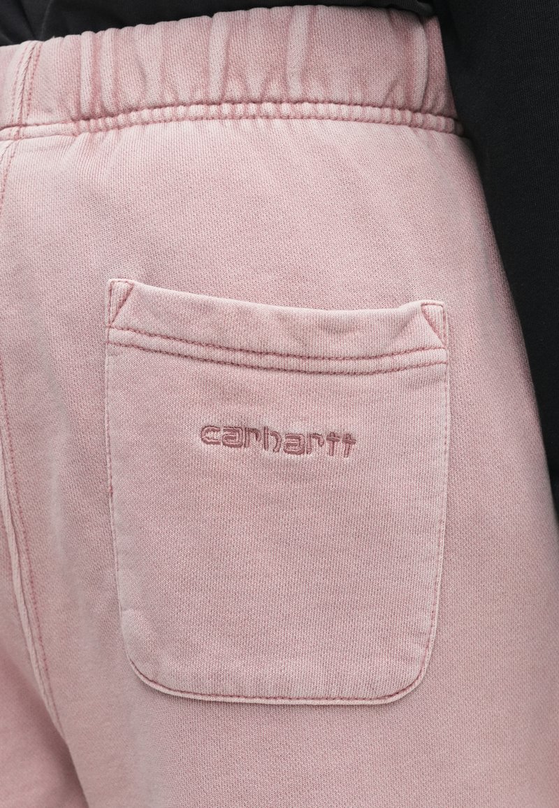 Pantaloni della tuta rosa con vita elastica e tasca posteriore ricamata con il logo Carhartt, indossati con un capo nero.