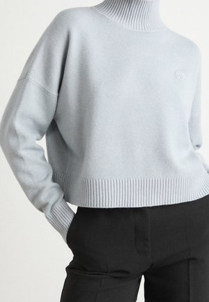 Pull à col roulé côtelé bleu clair à manches longues, associé à un pantalon noir taille haute, main dans la poche.