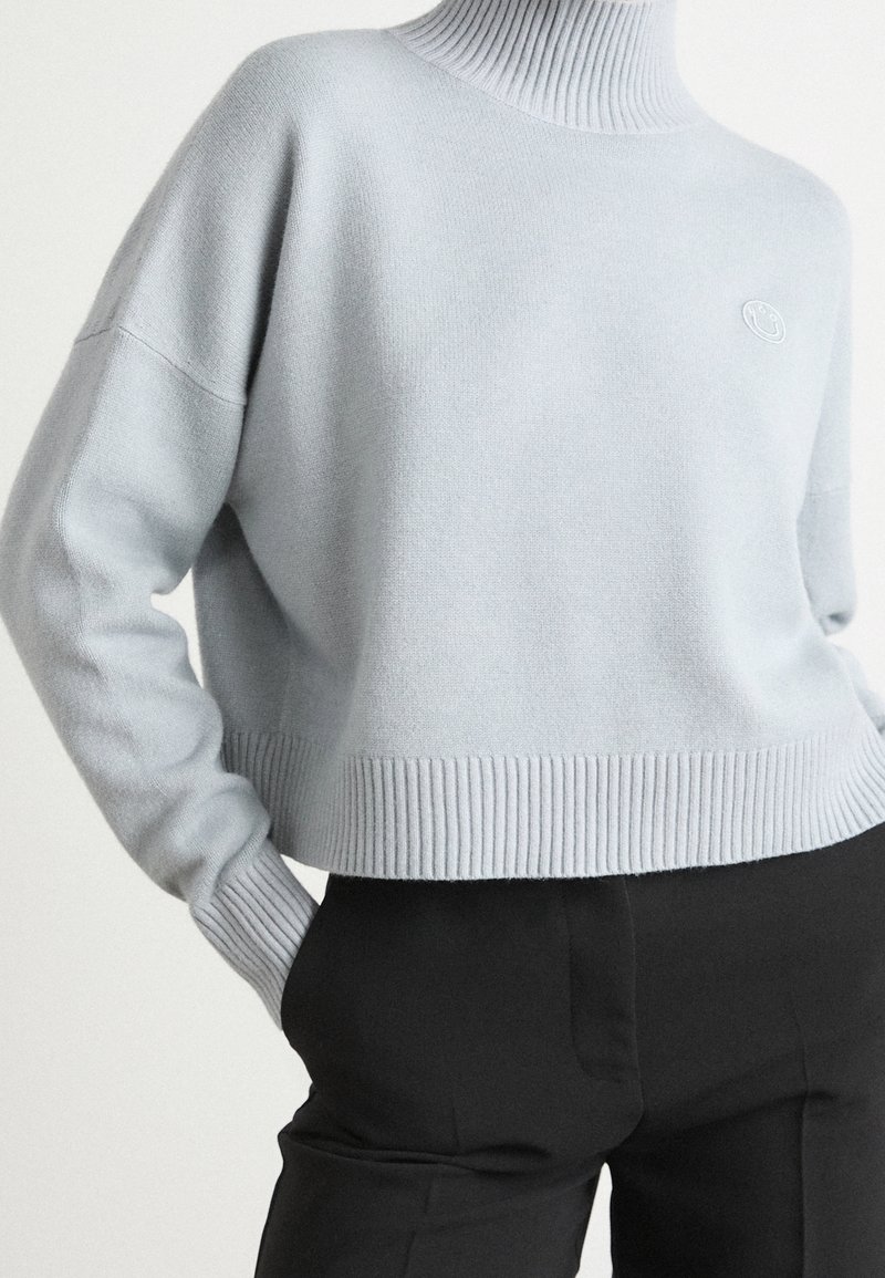 Pull à col roulé côtelé bleu clair à manches longues, associé à un pantalon noir taille haute, main dans la poche.
