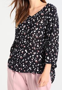 Schwarzbluse mit langen Ärmeln und V-Ausschnitt, versehen mit einem verstreuten Muster aus weißen und pinken Punkten sowie einem transparenten, leichten Stoff.