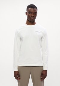 Bílá košile s dlouhým rukávem, kulatým výstřihem a detaily loga na hrudi, kombinovaná se světle hnědými kalhotami. Hladká struktura látky.