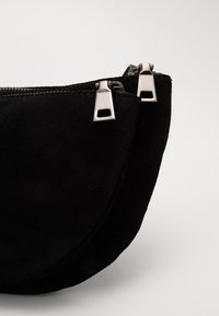 Černá semišová kabelka na ruku s prohnutým designem. Obsahuje dva stříbrné kovové zipy a hladký, texturovaný povrch.