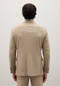 Giacca da uomo color beige chiaro realizzata in tessuto liscio, con uno spacco singolo, vestibilità su misura e bottoni decorativi sulle maniche. Vista posteriore mostrata.