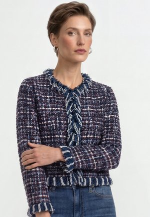 Woman with short hair wearing silver hoop earrings and a textured blue, white, and burgundy plaid jacket with fringed trim and blue jeans.