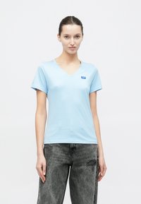 Ανοιχτό γαλάζιο μπλουζάκι με λαιμόκοψη σε σχήμα V, κατασκευασμένο από απαλό βαμβακερό ύφασμα, με κοντά μανίκια και μικρό μπλε λογότυπο στην αριστερή πλευρά του στήθους.