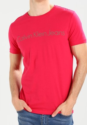 Mężczyzna w jasnoczerwonej koszulce Calvin Klein Jeans i niebieskich dżinsach z rękami w kieszeniach na jednolitym jasnym tle.