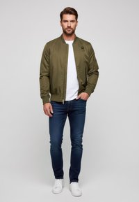 Giacca bomber verde oliva con polsini a coste, zip frontale, tasche laterali e una toppa. Indossata sopra una maglietta bianca e jeans blu scuro, con sneakers bianche.