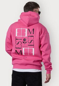 Haut à capuche rose avec impression graphique comprenant du texte et un design floral au dos, fabriqué en matériau doux, avec des poignets et un ourlet côtelés.