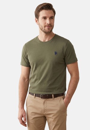 Man draagt olijfgroen T-shirt met korte mouwen met marineblauw logo en beige broek met donkerbruine riem, staande met één hand in de zak.