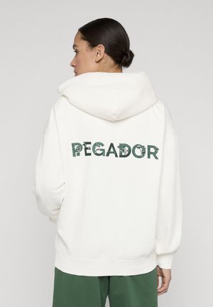 Kvinna med mörkt hår i knut som står i profil och bär en vit huvtröja med texten "PEGADOR" i grönt blommönster på ryggen.