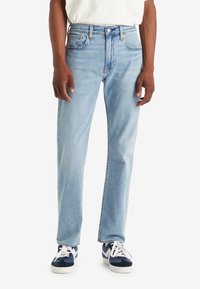 Personne portant un jean droit bleu clair, un t-shirt blanc et des baskets bleu marine à lacets blancs, debout devant un fond blanc uni.