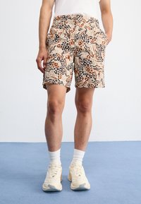 Pantaloni corti beige con un motivo astratto in marrone, nero e sabbia. Presentano tasche e una vestibilità casual. Indossati con sneaker e calzini di colore chiaro.