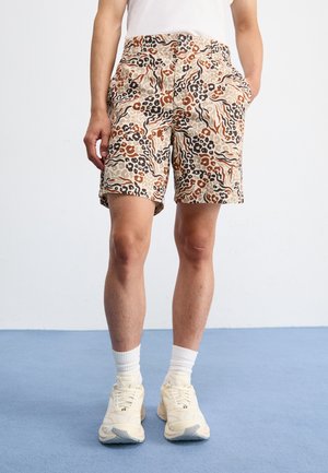 Pantaloni corti beige con un motivo astratto in marrone, nero e sabbia. Presentano tasche e una vestibilità casual. Indossati con sneaker e calzini di colore chiaro.