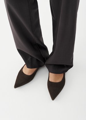 Fødder iført mørkebrune sko med spidse tæer og løse sorte bukser, der let dækker anklerne, stående på en hvid overflade.