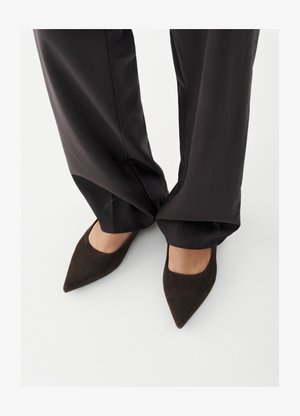 Pieds portant des chaussures à bout pointu brun foncé et un pantalon noir ample qui couvre légèrement les chevilles, debout sur une surface blanche.