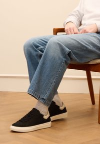 Black sneakers with a textured upper, white sole, and beige accents, paired with light blue cuffed jeans and grey socks.