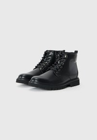 Botas de tobillo de cuero negro con un acabado liso, que cuentan con ojales de metal, parte delantera con cordones y una suela de goma texturizada. Incluye detalle de cremallera lateral.