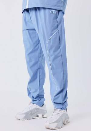 Persona que lleva pantalones deportivos de color azul claro con ribetes blancos y zapatillas Nike blancas frente a un fondo blanco liso.