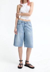 Bermudas de mezclilla azul claro con un ajuste relajado, dobladillo deshilachado y diseño de cinco bolsillos. Combinadas con una camiseta blanca sin mangas ajustada y sandalias negras.