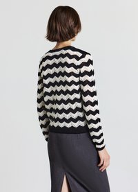 Pull en maille noir et blanc à motif zigzag avec un ourlet côtelé et des manches longues, fabriqué dans un matériau léger, porté par-dessus une jupe gris foncé.