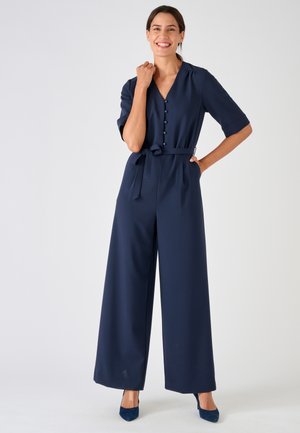 Donna sorridente, che indossa una tuta blu navy con bottoni, cintura annodata in vita e pantaloni a gamba larga, in piedi con una mano sul fianco.