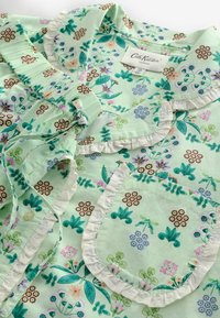 Vaaleanvihreä kukkakuosinen paita, jossa on röyhelökaulus ja tasku, ja jossa on vaaleanvihreitä ja tummanvihreitä lehtiä sekä pieniä värikkäitä kukkia.