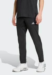Pantalons de jogger noirs avec des poignets élastiques, des détails texturés sur les côtés et un logo Adidas réfléchissant sur la cuisse gauche. Portés avec des baskets blanches.