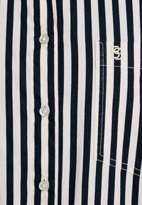 Camicia a righe con bottoni, caratterizzata da righe verticali blu navy e crema, bottoni bianchi e un piccolo logo ricamato sulla tasca del petto.