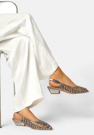 Benen in witte wijde broek, met zilveren gespen versierde slingback schoenen met lage blokhakken, de ene voet rustend op een stoeltoonladder en de andere omhoog.