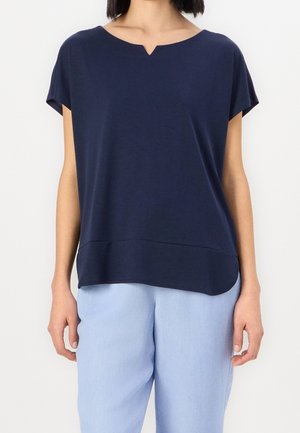 Femme portant un haut bleu marine à manches courtes avec une petite encolure en forme d'encoche et un pantalon ample bleu clair, sur un fond uni.