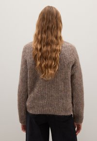 Een mokka-kleurige gebreide trui met een gestructureerd, ribbelpatroon. Losse pasvorm met lange mouwen, gedragen over donkere broek. Zacht, pluizig materiaal.