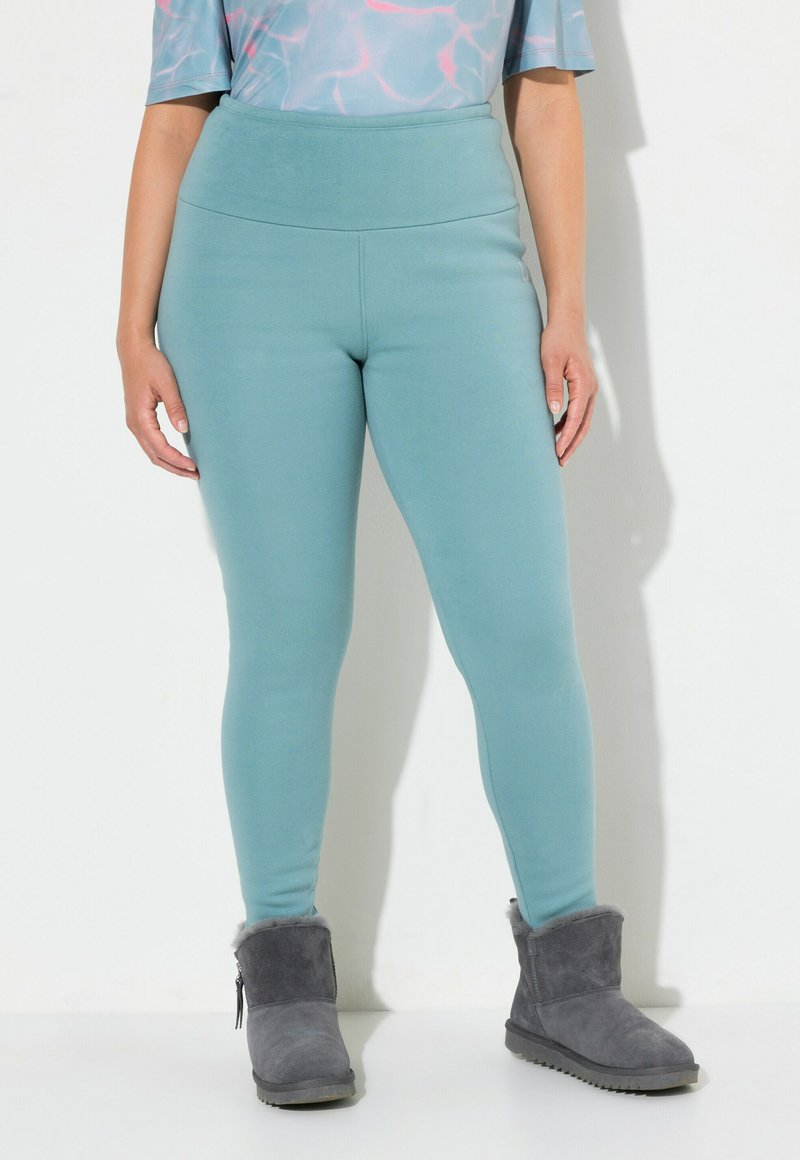 Donna in leggings teal a vita alta e stivali alla caviglia foderati in grigio, in piedi contro uno sfondo bianco semplice.
