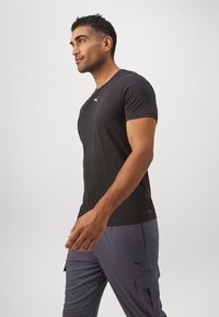 Svart tettsittende T-skjorte laget av mykt materiale, med en liten hvit Puma-logo på brystet, sammen med grå cargo-bukser.
