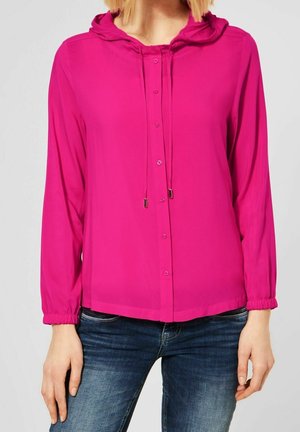 Persoon draagt een felroze capuchonblouse met knopen en trekkoorden en een blauwe spijkerbroek, staand tegen een effen lichte achtergrond.