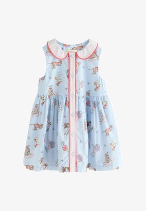 Mosolygó kislány copfokkal, állatmintás világos ruhában, kitárt karokkal áll egy színes virágpiac háttere előtt.