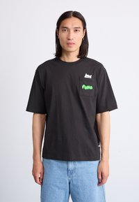 Black cotton t-shirt with short sleeves, featuring a white cat graphic on the chest pocket and green text reading "PUMA." Plain design, relaxed fit.
