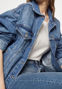 Chaqueta de mezclilla en azul claro con un corte holgado, gran cuello y dos bolsillos frontales. Presenta botones metálicos y un ligero detalle desgastado.
