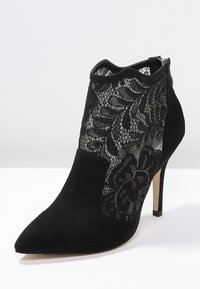 Stivaletto nero con tacco alto alla caviglia, in suede e pannelli di pizzo, punta affusolata e chiusura con zip. Presenta dettagli del design in pizzo floreale.
