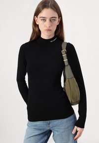 Svart langermet genser med høy hals i strikkematrealet, med en subtil logo ved kragen. Kombinert med en olivenfarget skulderveske.