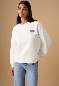 Hanorac alb cu textul „ECHOES OF FREEDOM” în negru, cu gât rotund, mâneci lungi și material moale texturat. Asortat cu blugi bleu deschis.