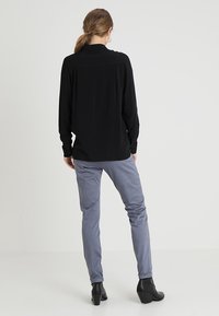 Schwarzes langärmliges Hemd mit einem Button-Down-Kragen, kombiniert mit hellgrauen Hosen und schwarzen Stiefeletten, von hinten betrachtet.