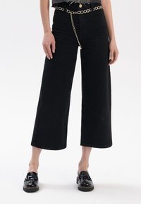 Zwarte denim wijde pijpen broek met een gemiddelde taille, voorzien van een rafelige zoom, zijsplitten en een zilveren kettingriem accent. Glanzende zwarte loafers maken de look compleet.