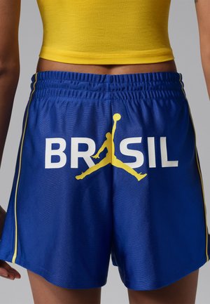 Személy kék atlétikai rövidnadrágot visel, melyen a „BRASIL” felirat és egy sárga kosárlabda jumpman logo található a hátsó részen, sárga crop toppal párosítva.