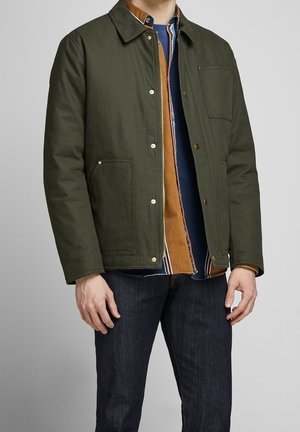 Mann trägt eine dunkelgrüne, geknöpfte Jacke über braunen und blauen geschichteten Hemden mit dunkelblauen Jeans, steht vor einem einfarbigen Hintergrund.