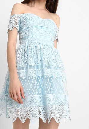 Robe en dentelle bleu clair épaules dénudées avec motifs complexes et ourlet festonné, portée par une personne aux longs cheveux bruns.