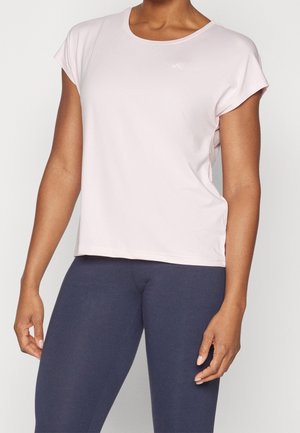 Kvinde iført en lyserød kortærmet skjorte og mørkeblå leggings stående mod en ensfarvet, lys baggrund.